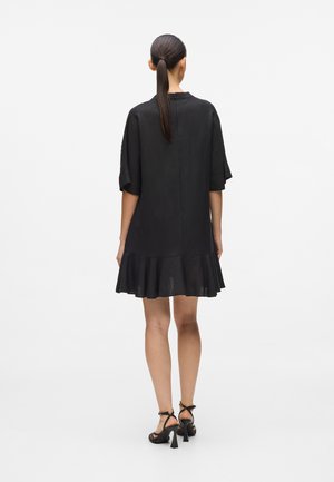 Frau von hinten, trägt ein kurzes schwarzes Kleid mit ausgestelltem Saum und Ellbogen-langen Ärmeln, kombiniert mit schwarzen, Riemchen-Sandaletten mit hohen Absätzen.