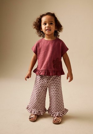 Kleinkind steht und trägt ein burgunderfarbenes kurzärmeliges Oberteil mit Rüschensaum und Blumenmotiv, gemusterte Hosen mit weitem Bein und roségoldene Sandalen.