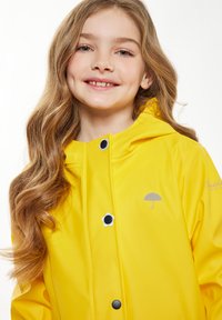 Geltonas nepralaidus lietpaltis su kapišonu, su dekoratyviniais juodais ir baltais gėlių mygtukais ir sidabro spalvos skėčio logotipu ant krūtinės.