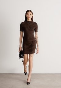 Γυναίκα με μακριά σκούρα μαλλιά που φοράει καφέ στενό φόρεμα με κοντά μανίκια, κρατώντας μία μικρή μαύρη τσάντα, περπατά σε γκρι πάτωμα μπροστά από λευκό τοίχο.