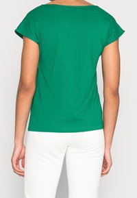 Person i grön kortärmad topp och vita byxor, stående med avslappnade armar, med ryggen mot betraktaren framför en enkel bakgrund.