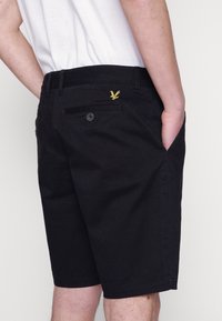 Sorte shorts lavet af stof med sidelommer, en baglomme med knap og et lille gult broderet logo på den bagerste talje.