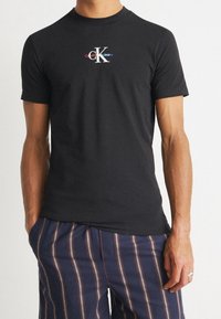 Svart crew neck t-shirt i bomull med en framsida som har en grafik med "Calvin Klein Jeans" och en stiliserad 'K' i vitt, rött och blått.