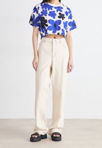Camisa blanca recortada con patrones de flores azules y negras, combinada con jeans de tiro alto en color crema y sandalias de plataforma negras.