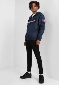 Windbreaker azul-marinho com um bolso frontal em formato de bolsa e detalhe de logotipo, combinado com calças pretas justas e ténis pretos. Material suave e leve.