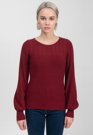 Burgunderfarbener gestrickter Pullover mit einem strukturierten Muster am Ausschnitt, langen Puffärmel und gerippten Bündchen. Kombiniert mit dunkelblauen Jeans.