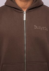 Smeđa dukserica na zip, izrađena od pamuka, s kapuljačom, koja ima srebrni patentni zatvarač i suptilan izvezeni logo na lijevoj strani prsnog koša.