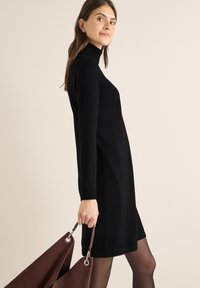 Schwarzes Rollkragenkleid mit langen Ärmeln, lockerem Schnitt und Seitenschlitzen. Das Model hält eine dunkelbraune Wildledertasche mit silbernen Beschlägen.