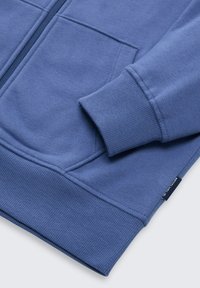 Blauer Sweatshirt mit gerippten Bündchen, Reißverschluss und seitlichen Taschen. Weicher Stoff mit glatter Textur. Verfügt über ein kleines Markenetikett.