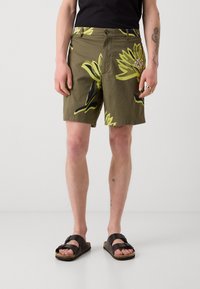 Personne portant un short vert olive avec un grand imprimé floral jaune, un T-shirt noir, des sandales noires et des tatouages visibles sur les jambes.