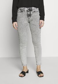 Jeans ajustados en denim gris desgastado con una ligera textura, con bolsillos frontales, lazos para cinturón y cierre de botón. Combinados con sandalias deslizantes negras.