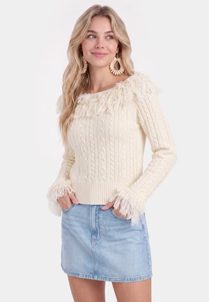 Jonge vrouw die een crèmekleurige kabelgebreide trui met franjes draagt en een lichtblauwe spijkerbroekrok, handen in de zakken, licht glimlachend naar rechts kijkend.