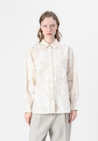 Blusa trasparente crema con intricati ricami floreali bianchi, chiusura frontale con bottoni, colletto e maniche lunghe. Tessuto morbido, vestibilità comoda.