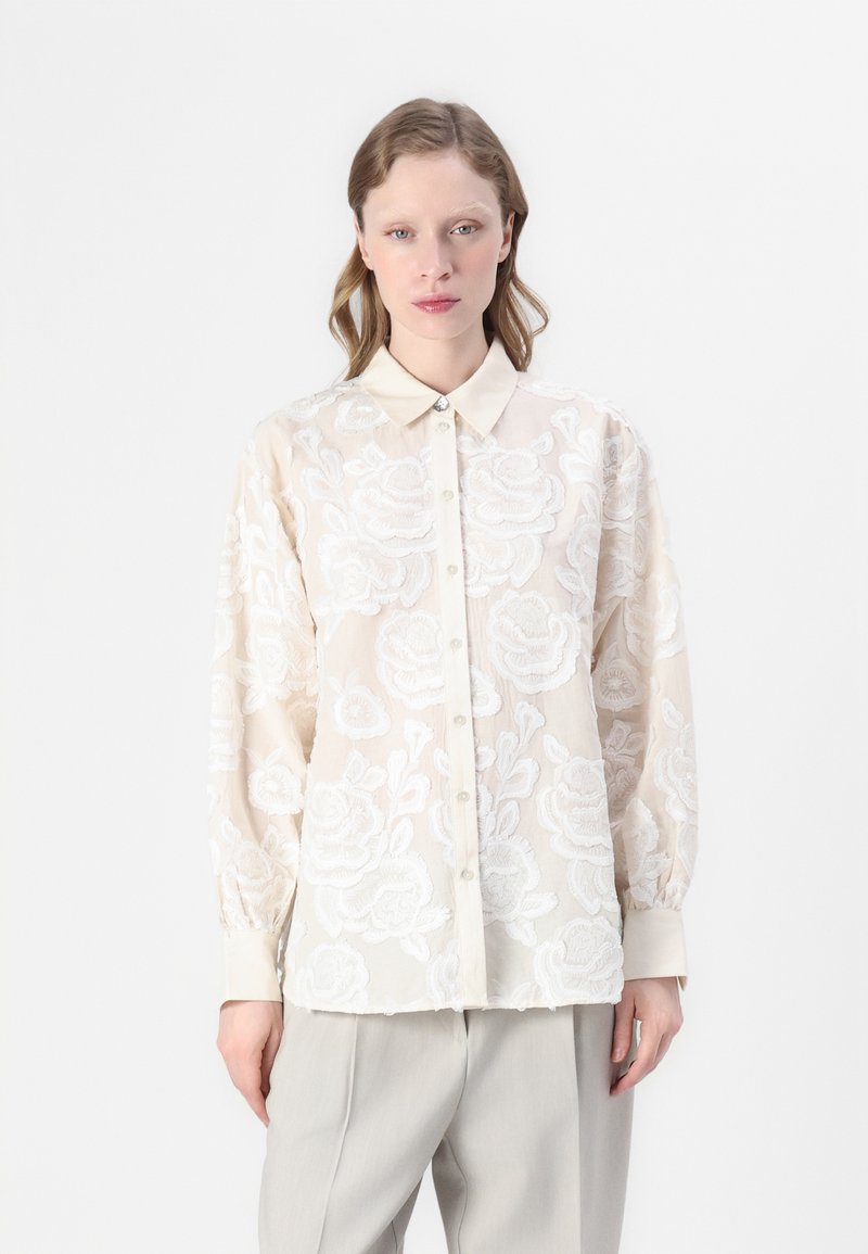 Blusa trasparente crema con intricati ricami floreali bianchi, chiusura frontale con bottoni, colletto e maniche lunghe. Tessuto morbido, vestibilità comoda.