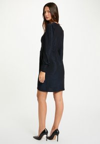 Femme aux longs cheveux bruns portant une mini-robe à manches longues bleu foncé et des talons hauts noirs, se tenant debout et regardant par-dessus son épaule.