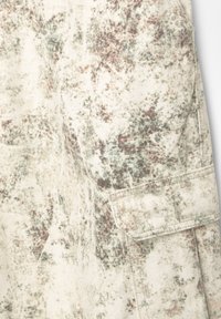 Nærbillede af stof med et beige, grønt og brunt camouflage-mønster, der har en syet rektangulær lomme med klap.