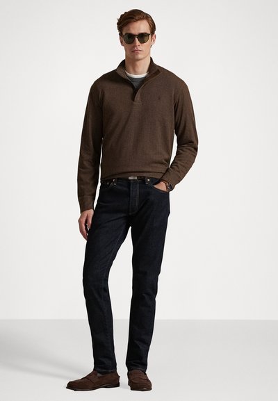 Καφέ, υφασμάτινη μπλούζα με φερμουάρ μέχρι το 1/4, συνδυασμένη με σκούρα τζιν και καφέ λουστρίνια. Περιλαμβάνει γυαλιά ηλίου και χαλαρή στάση.