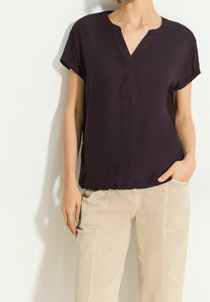 Kvinde iført en sort bluse med korte ærmer og V-hals samt beige bukser, stående med den ene hånd i lommen mod en ensfarvet baggrund.