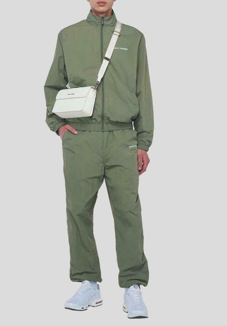 Olivgrüner Trainingsanzug mit weißer Markenkennung, leichtes Material, Jacke mit Reißverschluss, elastischer Taillenbund. Weiße Umhängetasche und Sneaker.