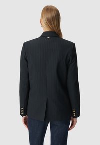 Een persoon met lang blond haar staat met de rug naar de camera, gekleed in een zwart gestreept colbert met gouden knopen en een blauwe spijkerbroek, tegen een effen achtergrond.