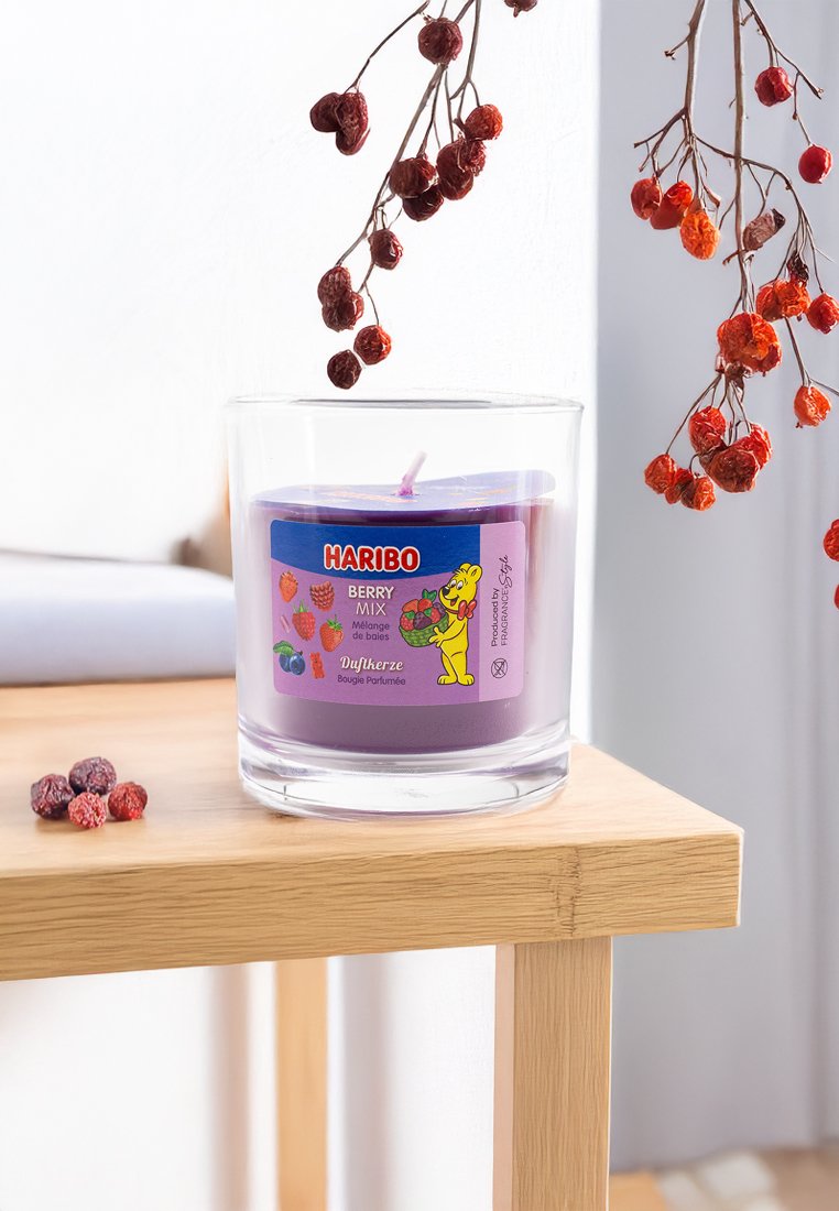 Vela aromática Haribo Berry Mix morada en un tarro de cristal sobre una mesa de madera, con bayas rojas secas colgando arriba y esparcidas cerca.