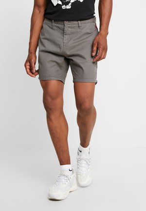 Modèle masculin portant un short gris mi-cuisses, des baskets blanches et un t-shirt noir avec un motif, debout devant un fond blanc uni.