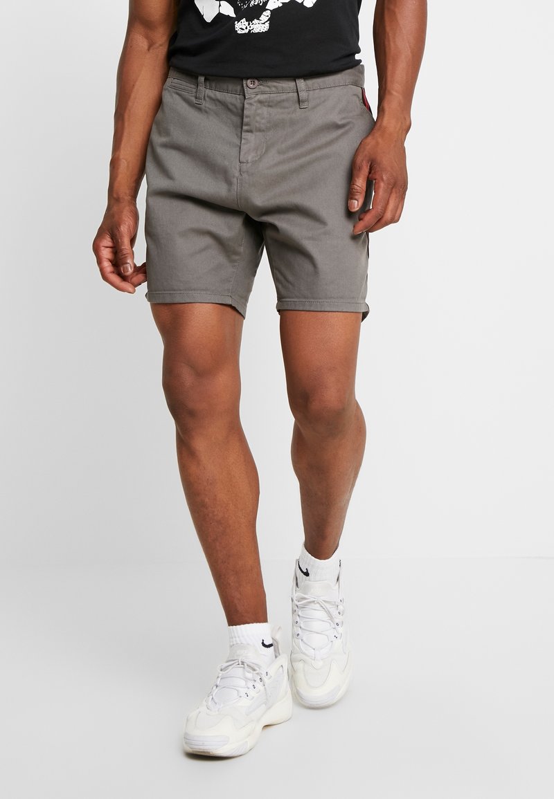 Modèle masculin portant un short gris mi-cuisses, des baskets blanches et un t-shirt noir avec un motif, debout devant un fond blanc uni.