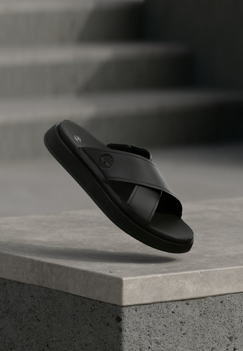 Μαύρο δερμάτινο σανδάλι με σταυρωτούς ιμάντες και λογότυπο δέντρου να αιωρείται πάνω από μια επιφάνεια από μπετόν με θολωμένα σκαλοπάτια στο φόντο.