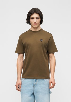 Mladý muž s vlasmi po plecia, oblečený v olivovo zelenej tričku s krátkym rukávom a svetlo modrých džínsoch, stojaci pred jednoduchým pozadím.