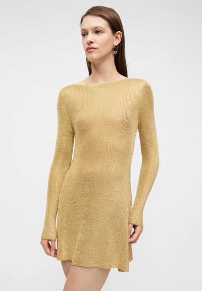 Γυναίκα με ίσια καστανά μαλλιά, φοράει χρυσλό πλεκτό φόρεμα με μακριά μανίκια και χρυσούς κρίκους σκουλαρίκια, κοιτάζει ελαφρώς προς τα δεξιά σε απλό φόντο.