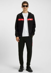Ung mand iført sorte solbriller, sort zip-up jakke med røde striber, hvid T-shirt, sorte bukser og sorte sneakers, stående mod en ensfarvet baggrund.