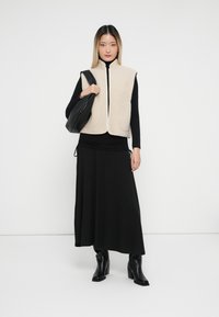 Žena v čiernom roláku, čiernej dlhé sukni, béžovej bezrukávovej zipovej veste, čiernych čižmách, drží čiernu ramennú tašku na bielom pozadí.
