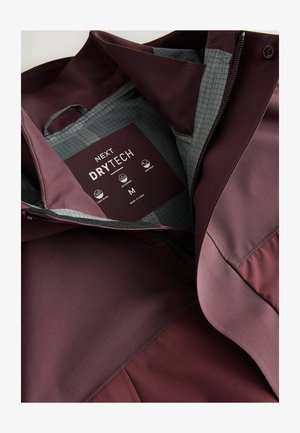 Maroon waterproof jacket with grey grid-patterned lining, label showing "NEXT DRYTECH," size M, windproof and taped seams features.
