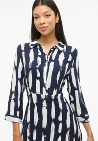 Jūrinė marškinėlių suknelė su balta teptuko štrichų raštu, mygtukų užsegimu priekyje ir ilgomis rankovėmis su rankogaliais. Minkšta medžiaga su prigludusia juosmens dalimi.