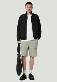 Casaco preto com fecho éclair sobre uma t-shirt branca, combinado com calções cinzentos claros. Ténis pretos e uma bolsa preta. Tecido liso, design casual.