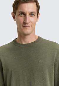 Zielony sweter dzianinowy z okrągłym dekoltem i ściągaczami na rękawach. Na piersi subtelne logo. Gładka faktura i klasyczny design.