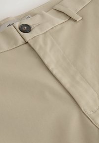 Primer plano de la cintura de unos pantalones beige con botón negro, presilla para el cinturón y costuras visibles cerca de la bragueta.