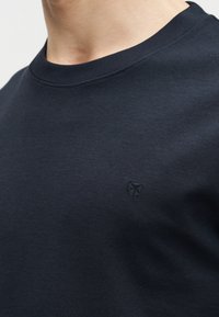 Κοντινή εικόνα ενός ατόμου που φοράει σκούρο μπλε φούτερ με λαιμόκοψη, με ένα μικρό, διακριτικό κεντητό λογότυπο στο αριστερό μέρος του στήθους.