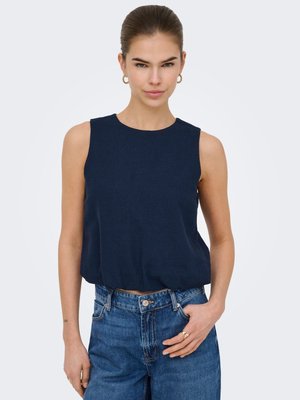 Mujer con el cabello recogido, lleva pendientes de aro dorados, una blusa sin mangas azul marino y vaqueros azules, con la mano en el bolsillo.