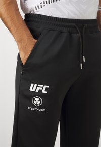 Čierne športové nohavice s elastickým pásom, zdobené bielymi logami "UFC" a "crypto.com". Obsahujú bočnú zipsovú kapsu.