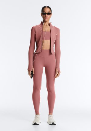 Femeie în picioare, purtând haine sport roz, cu jachetă și colanți asortați, pantofi sport albi, ochelari de soare și căști, ținând un telefon.