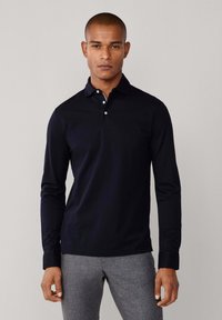 Mornarska polo majica dugih rukava s ovratnikom, s plastičnim kopčanjem na tri gumba i glatkom teksturom tkanine. Uparena s sivim hlačama.