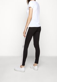Leggings neri con una striscia di logo bianca lungo il lato, abbinati a una t-shirt bianca aderente e scarpe da ginnastica bianche.