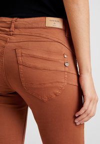 Jeans en denim de couleur rouille avec une coupe ajustée. Comprend une étiquette de logo crème, deux poches arrière et des détails en métal argenté.