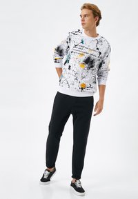 Vit sweatshirt med färgglad splatter-mönster och text, kombinerad med svarta joggers. Sneakers har svart tyg och vita sulor.