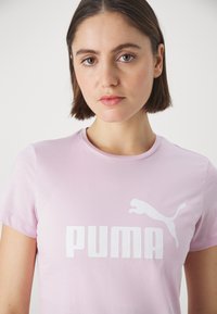 Svetloružové tričko z hladkej bavlny s bielym logom "PUMA" a grafikou skáčucej mačky na prednej strane. Krátke rukávy a okrúhly výstrih.