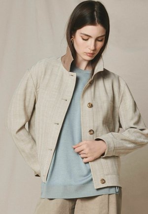 Frau trägt eine beige Fischgrätjacke mit hohem Kragen über einem hellblauen Pullover und richtet die Jackenvorderseite mit einer Hand.