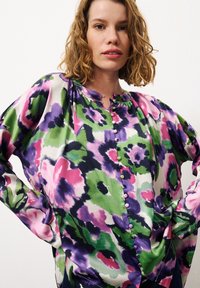 Bluse mit floralem Muster in lila, grün und pink. Seidiger Stoff, runder Ausschnitt, lange Ärmel und Knopfdetails auf der Vorderseite.