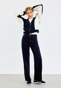 Skraćena navy i krem zip-up dukserica s oversized rukavima, uparena s crnim trapericama širokih nogavica i navy tenisicama s bijelim trakama.