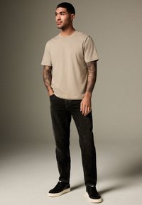 Mann mit Tattoos steht und trägt ein beiges T-Shirt, schwarze Jeans und schwarze Turnschuhe vor neutralem einfarbigem Hintergrund.